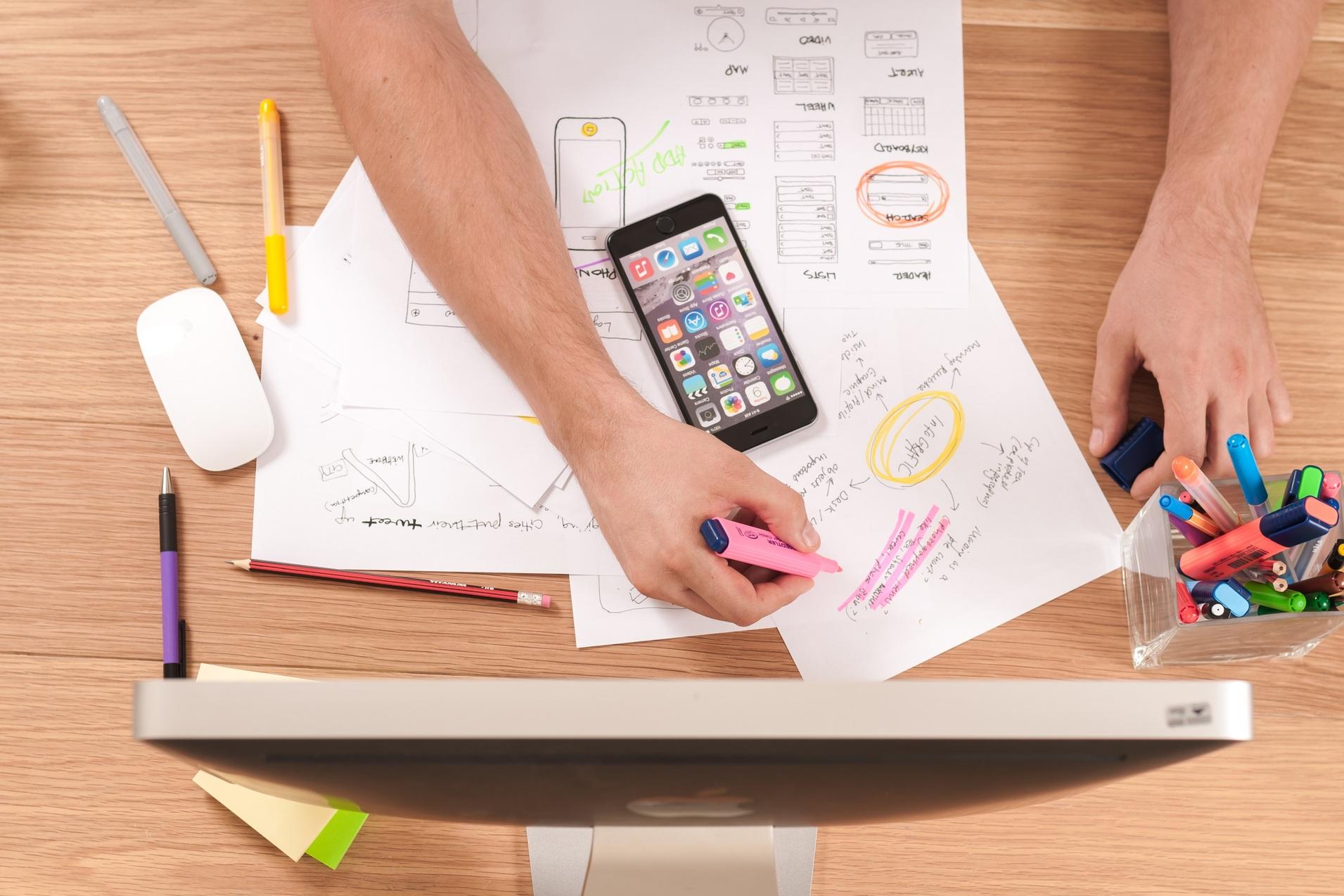Skizze und Entwicklung Hände mit Stiften und einem Smartphone auf einem Tisch, umgeben von Notizen und einem Computer.